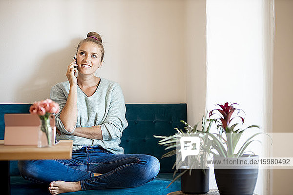 Porträt einer lächelnden blonden Frau am Telefon  die barfuß auf einer Bank zu Hause sitzt