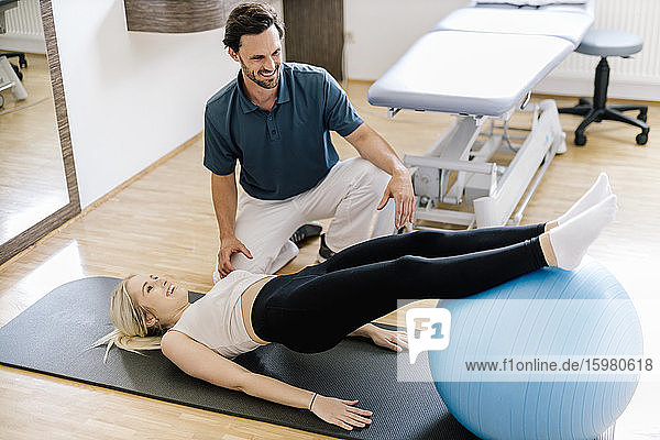 Physiotherapeutin hilft einer Patientin  die mit einem Fitnessball übt  auf einer Gymnastikmatte liegend