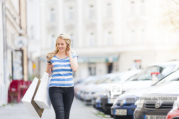 Porträt einer glücklichen blonden Frau mit Einkaufstüten  die auf ihr Smartphone schaut  während sie auf dem Bürgersteig geht