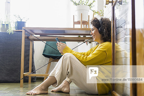 Lächelnde Frau  die zu Hause auf dem Boden sitzt  Kopfhörer trägt und ein Smartphone benutzt