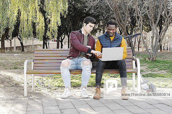 Two young men sitting on park bench sharing laptop