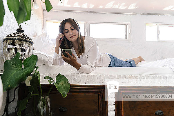 Porträt einer auf dem Bett liegenden Frau  die mit Kopfhörern Musik hört und auf ihr Handy schaut