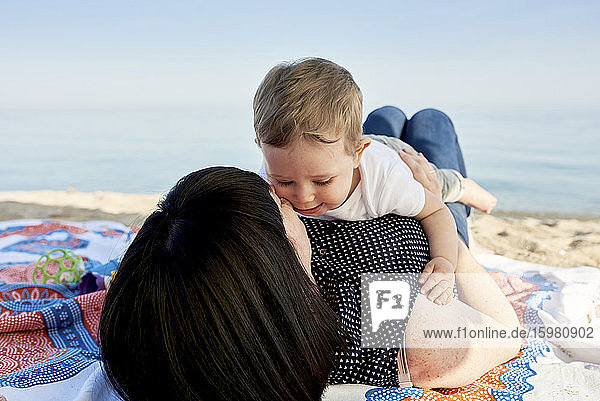 Mother kissing cute son while lying on blanket at beach against sky