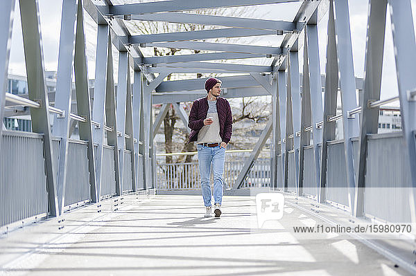 Junger Mann  der mit Ohrstöpseln und Smartphone auf einer Brücke spazieren geht