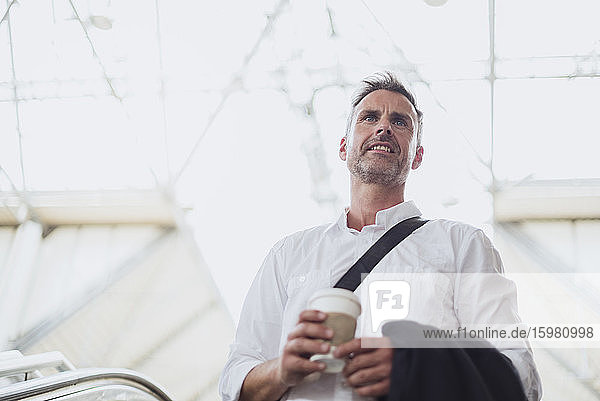 Nachdenklicher Geschäftsmann mit Kaffee in der Hand  der wegschaut  während er auf einer Rolltreppe steht