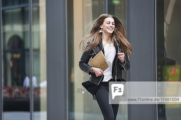 Cheerful young woman holding laptop while running in city
