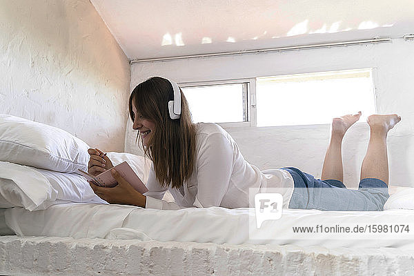 Smiling woman lying on bed listening music with headphones while taking notes