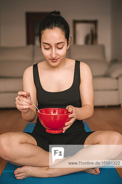 Junge Frau hält eine Frühstücksschüssel  während sie zu Hause auf einer Übungsmatte sitzt