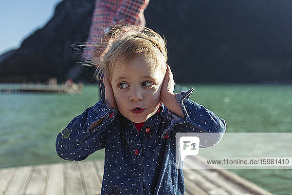Niedliches Mädchen  das sich die Ohren zuhält  während der Vater im Hintergrund auf der Strandpromenade am Achensee  Bundesland Tirol  Österreich  steht