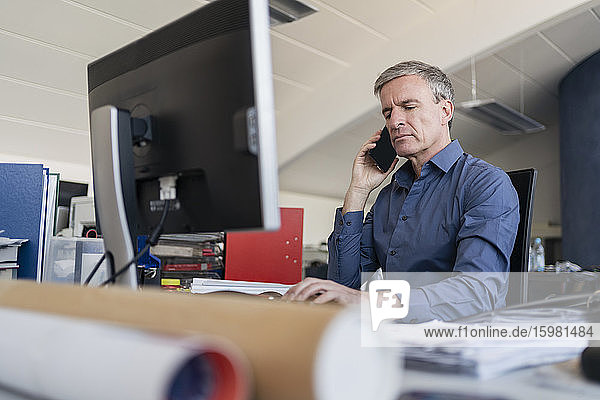 Handsome male professional talking on mobile phone while sitting at computer desk in office