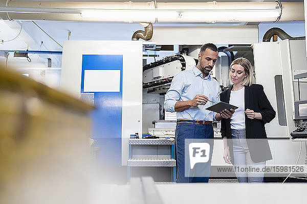 Geschäftsmann mit Tablet und Frau bei einer Besprechung an einer Maschine in einer Fabrikhalle