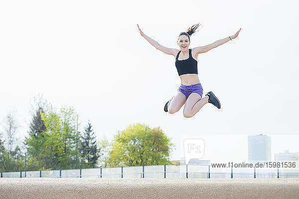 Cheerful young woman with arms outstretched jumping over land against clear sky