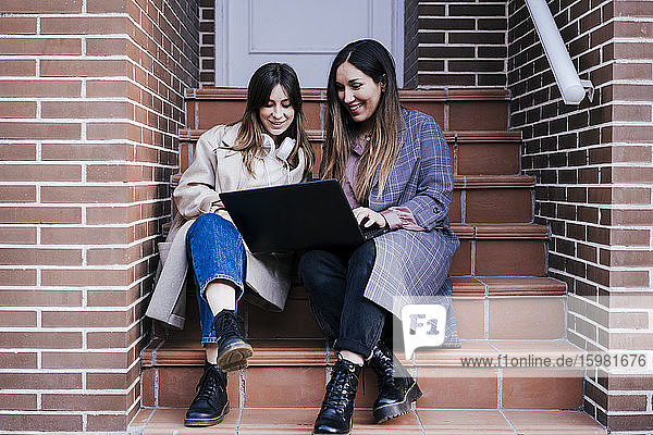 Porträt von zwei Frauen  die auf einer Treppe im Freien sitzen und auf einen Laptop schauen