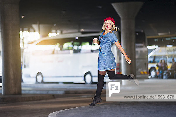 Porträt einer glücklichen jungen Frau mit Kaffee zum Mitnehmen  die am Busbahnhof in die Luft springt