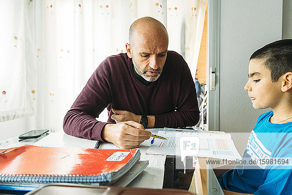 Confident father teaching son while sitting at desk during homeschooling