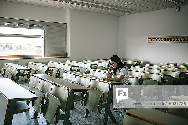 Full length of young pensive female student sitting at desk in university classroom