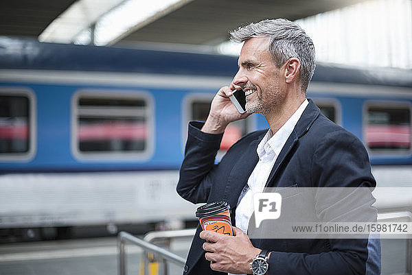 Geschäftsmann mit Kaffee in der Hand  der über sein Smartphone spricht  während er am Bahnhof steht
