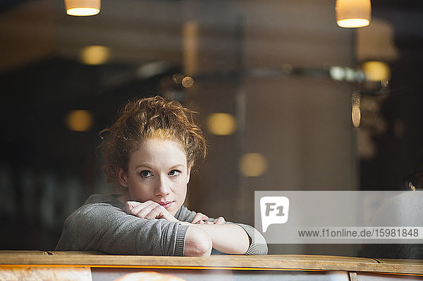 Nachdenkliche junge Frau  die sich auf einen Tisch stützt  gesehen durch ein Glasfenster in einem Café