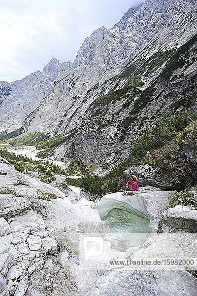 Ältere weibliche Touristin sitzt auf einem Felsen am Bach vor den Bergen