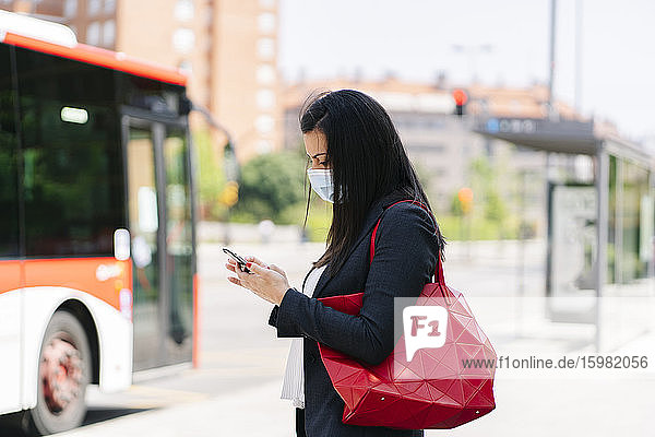 Frau mit Schutzmaske benutzt ihr Smartphone beim Warten an einer Bushaltestelle  Spanien