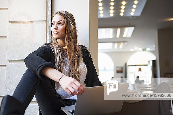 Nachdenkliche junge Frau sitzt mit Laptop auf der Fensterbank eines Cafés
