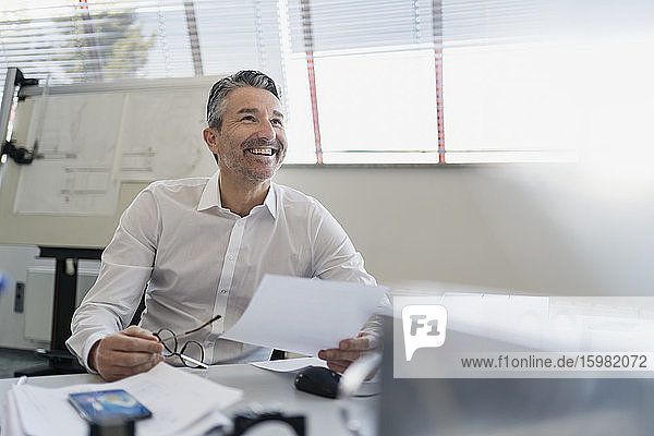 Happy handsome businessman looking up while sitting with paper at desk in office
