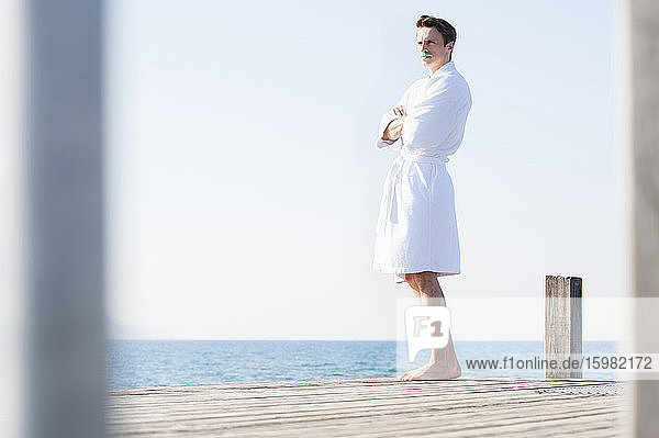 Gut aussehender Mann im Bademantel  der auf einem Pier über dem Meer vor einem klaren Himmel steht