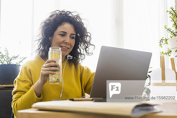 Woman using laptop at desk in home office drinking detox water