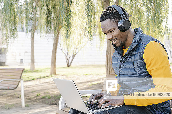 Junger Mann mit Kopfhörern sitzt auf einer Parkbank und benutzt einen Laptop