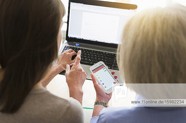 Rückenansicht einer älteren Frau und ihrer erwachsenen Tochter bei der Nutzung von Laptop und Smartphone