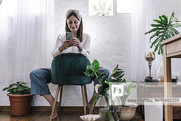 Lächelnde Frau  die ihr Smartphone benutzt  während sie auf einem Stuhl an der Wand zu Hause sitzt