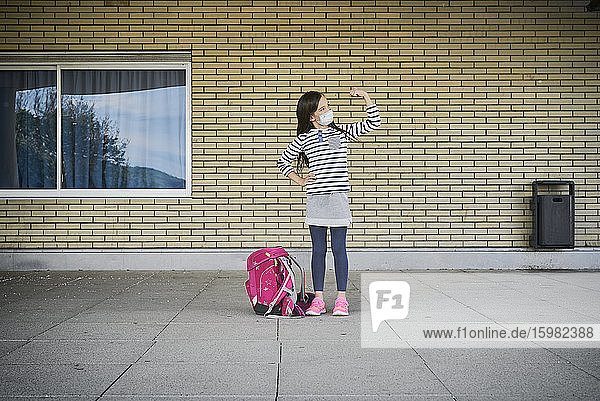 Mädchen mit Schultasche und Maske steht vor einem Gebäude und lässt ihre Muskeln spielen