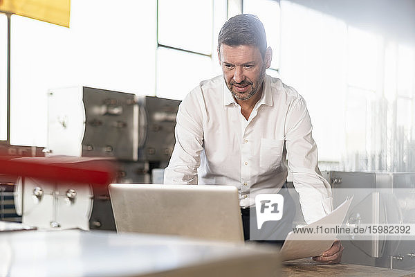 Älterer Geschäftsmann hält Papiere und benutzt einen Laptop in einer Fabrik