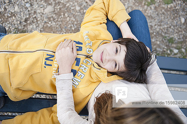 Lächelnder Junge auf dem Schoß der Mutter auf einer Parkbank liegend