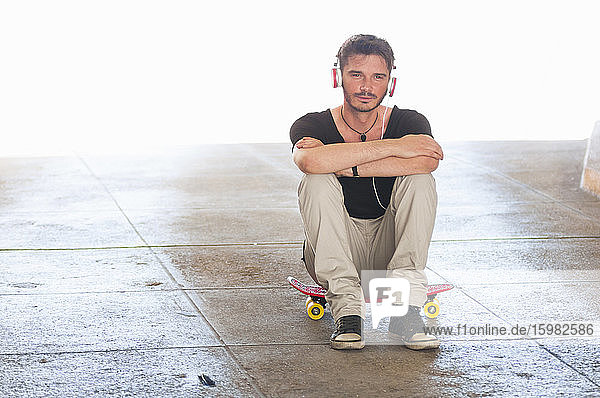 Portrait of man sitting on skateboard listening music with headphones