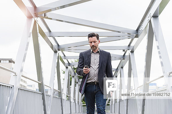 Confident handsome businessman using smart phone while walking on footbridge in city