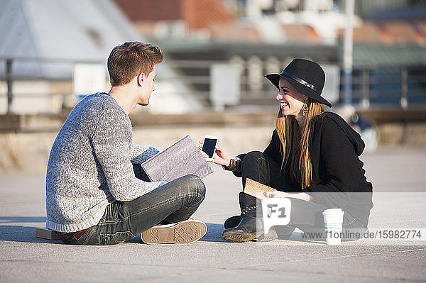 Happy couple holding smart phone and laptop while sitting on building terrace in city