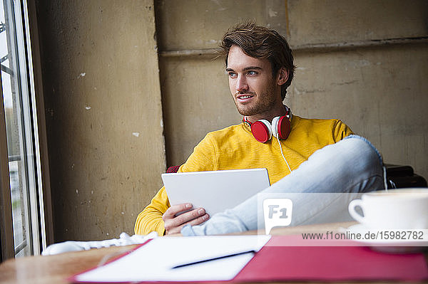 Porträt eines jungen Mannes mit Kopfhörern und digitalem Tablet in einem Café  der aus dem Fenster schaut