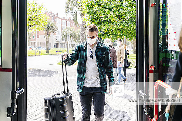 Junger Mann mit Schutzmaske steigt mit Trolley-Tasche in einen Bus ein  Spanien