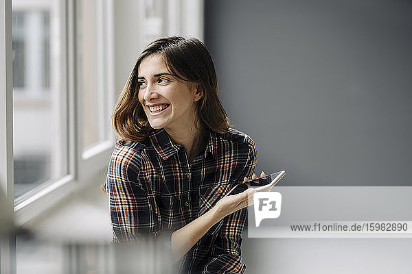 Porträt einer lachenden jungen Frau mit Smartphone  die aus dem Fenster schaut