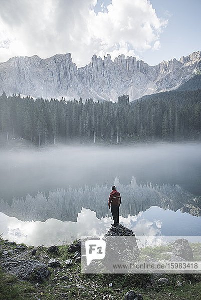 Italy  Carezza  Young man standing on rock at Lago di Carezza in Dolomite Alps at dawn