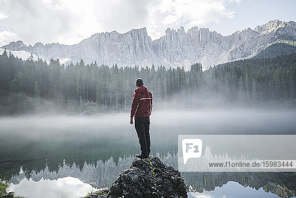 Italien  Carezza  Junger Mann steht auf Fels und schaut in der Morgendämmerung auf den Lago di Carezza in den Dolomiten