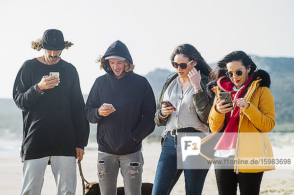 Freunde benutzen Mobiltelefone  während sie mit ihrem Hund im Sommer am Strand von Tarifa  Spanien  stehen