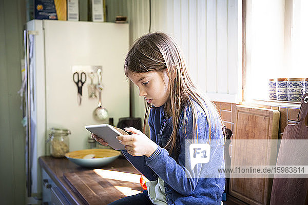 Mädchen sitzt auf der Arbeitsplatte in der Küche und schaut auf ein digitales Tablet