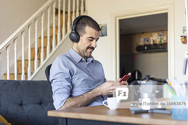 Mann arbeitet von zu Hause aus und benutzt in der Kaffeepause sein Smartphone mit Kopfhörern