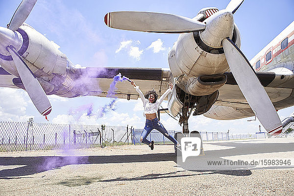 Unbeschwerte junge Frau genießt  während sie eine Notfackel mit lila Rauch gegen ein Flugzeug auf der Startbahn eines Flughafens hält
