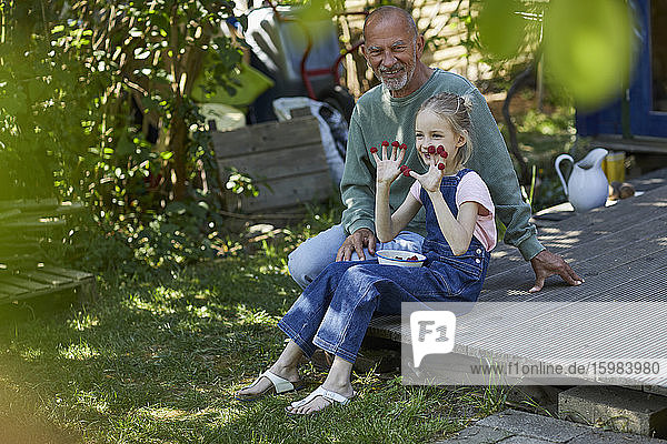Großvater und Enkelin mit Himbeeren auf ihren Fingerspitzen auf der Terrasse im Garten sitzend