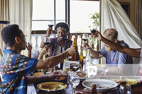 Lächelnde männliche und weibliche Freunde stoßen mit Getränken an  während sie während des Mittagessens am Esstisch sitzen