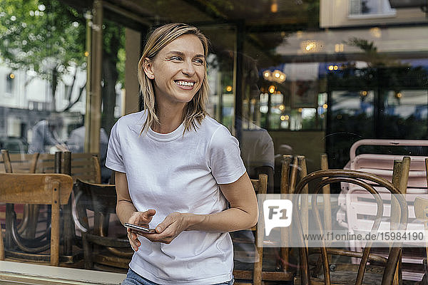 Porträt einer lächelnden Frau mit Smartphone  die vor einem geschlossenen Café steht und in die Ferne schaut