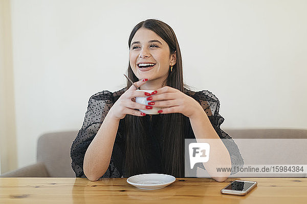 Fröhliche junge Frau hält eine Kaffeetasse  während sie am Tisch sitzt und in der Cafeteria wegschaut
